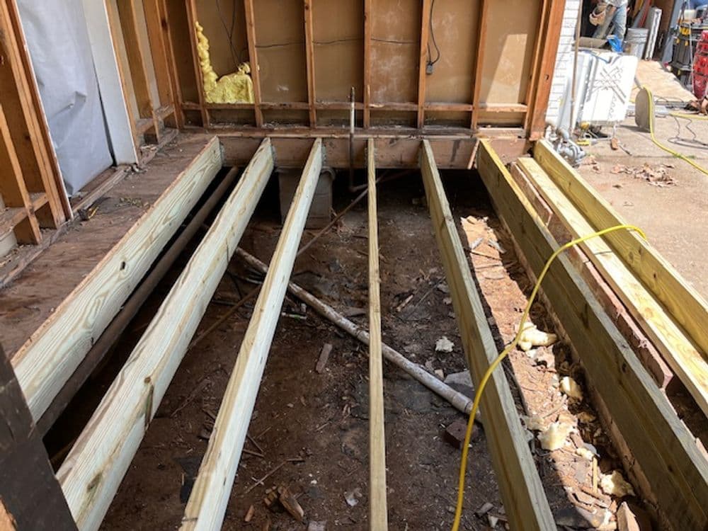 Wooden beams support a house foundation during renovation, with exposed framing and tools visible.