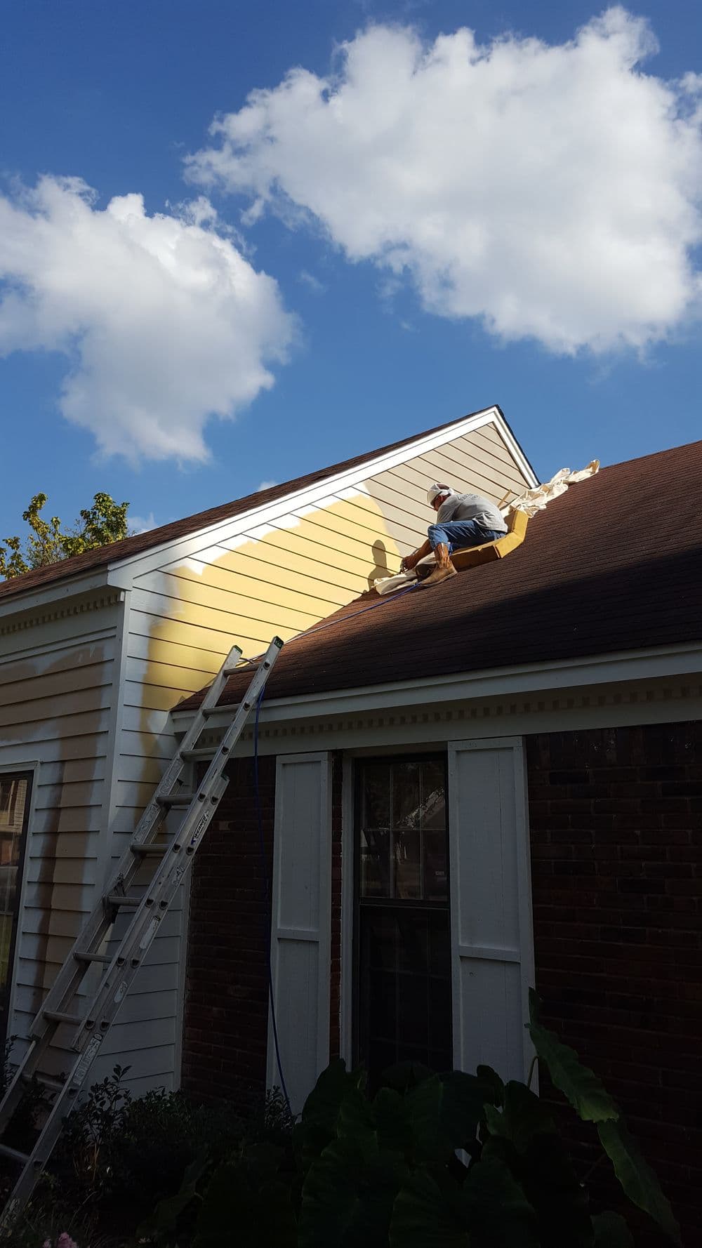 Person painting the exterior of a house while using a ladder against a sunny sky.