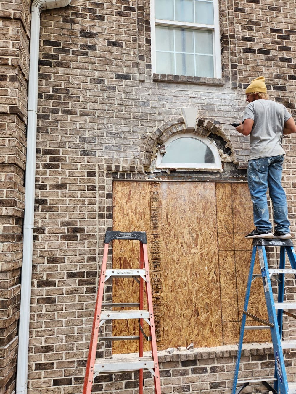 Construction worker repairing a brick wall with plywood boarding and ladders.