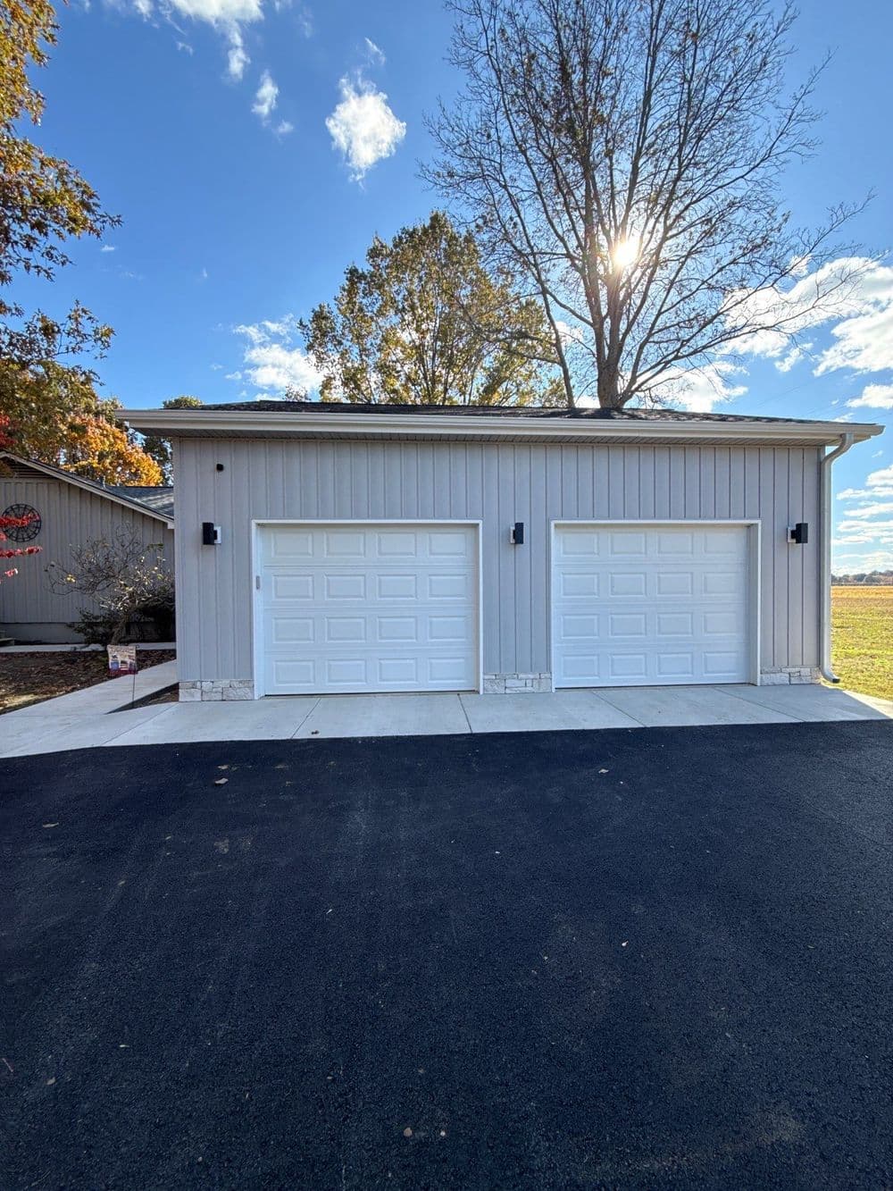 Modern two-car garage with white doors, asphalt driveway, and autumn trees under a blue sky.