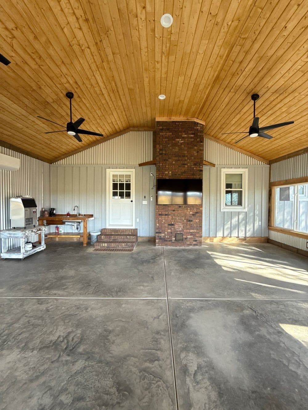 Spacious indoor outdoor living area with wood ceiling, brick fireplace, and modern kitchen setup.